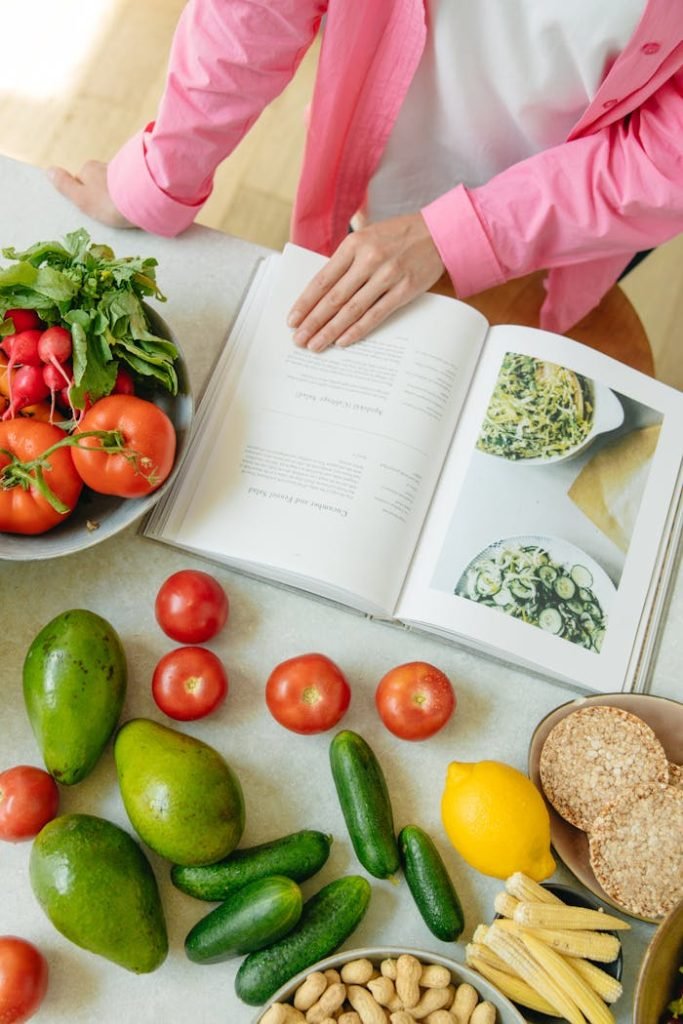 hand-of-a-person-on-an-open-book-near-fresh-vegetables-8845419 Overhead view of fresh vegetables and an open cookbook on a kitchen counter.