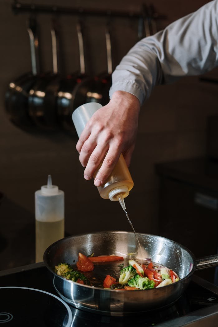 A chefs hand drizzling oil over mixed vegetables in a frying pan in a kitchen setting.